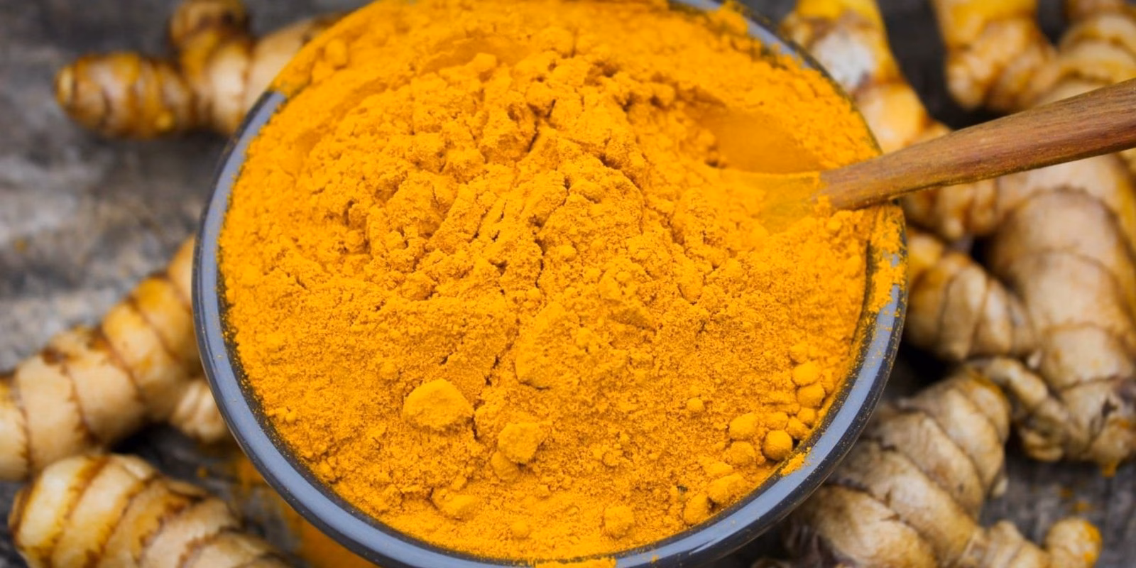 Cuenco de cerámica lleno de polvo de cúrcuma anaranjado con una cuchara de madera, rodeado de rizomas frescos de Curcuma longa sobre una superficie de piedra